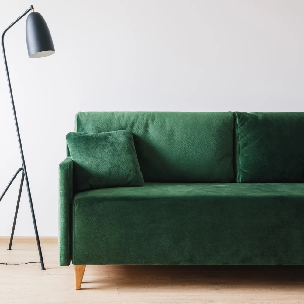 A green sofa is shown with a tall lamp next to it against a white wall and on wooden flooring.