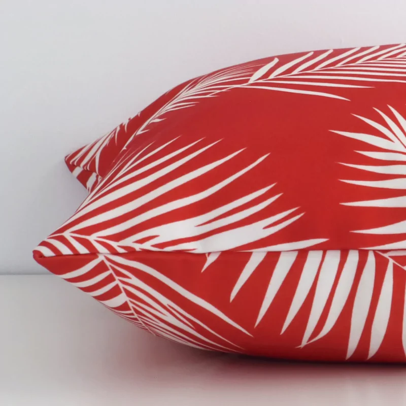 A red palm styled cushion arranged sideways in front of a wall. The square shape and outdoor fabric are shown and the seams are clearly visible.