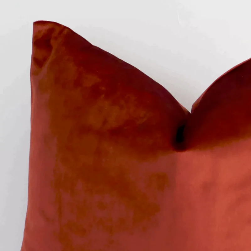 Macro shot showing the top left side of this square velvet cushion. The rust hue and solid-coloured design are magnified.