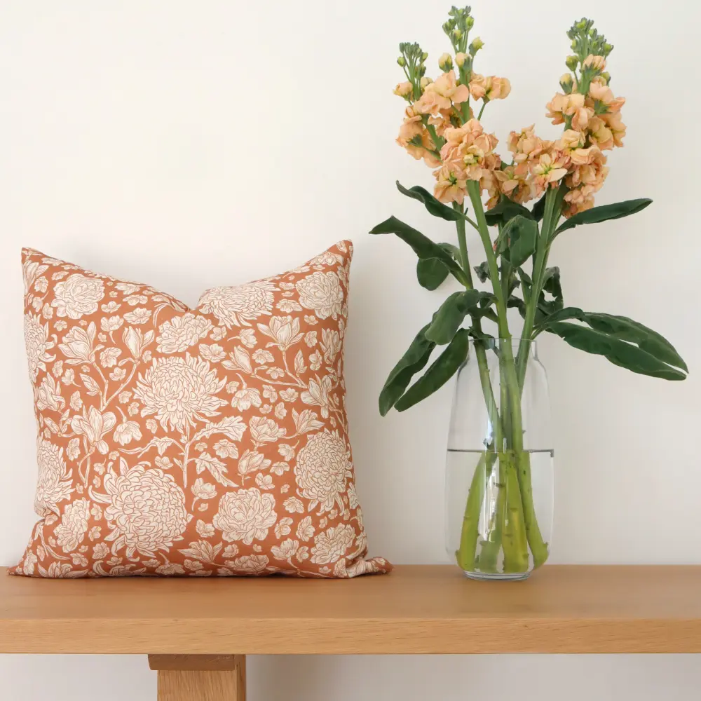 Floral cushions beside a glass vase with flowers in it all resting on a bench.
