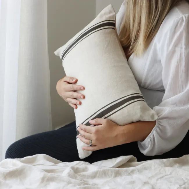 A lady is holding an elegant striped cushion with a curtain on the background.