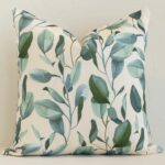Floral cushion in cream and green colour positioned in front of a brick wall. It has square dimensions and is made from an outdoor material.