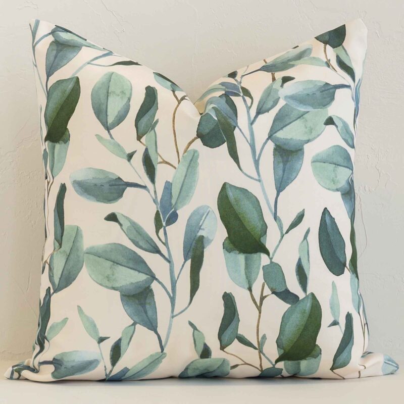 Floral cushion in cream and green colour positioned in front of a brick wall. It has square dimensions and is made from an outdoor material.
