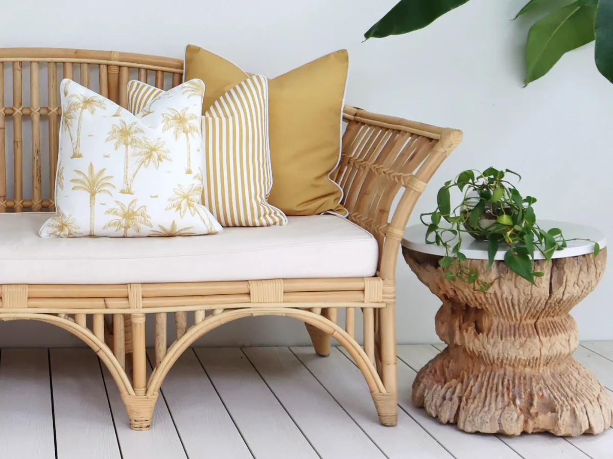 An outdoor couch is shown with 3 mustard outdoor cushions arranged to one side next to a side table with background foliage.