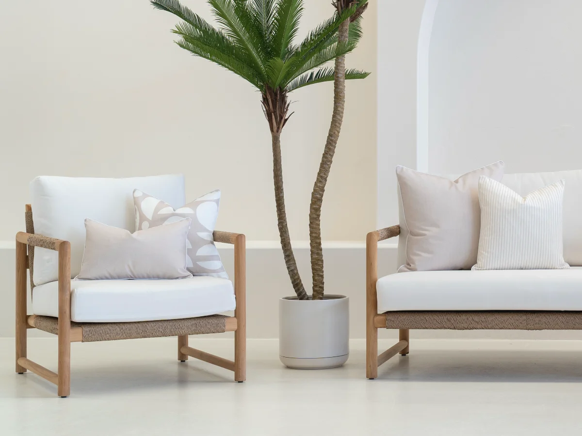 Sand beige outdoor cushions arranged on an outdoor armchair and sofa with a potted plant between them.